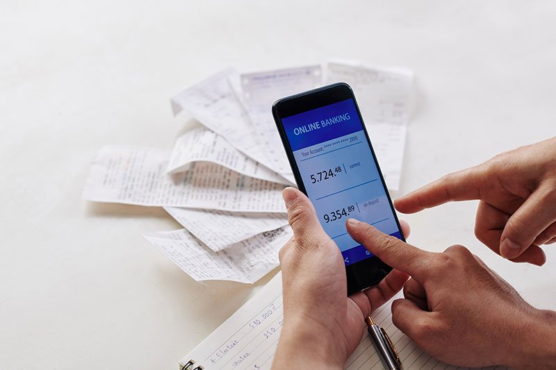 Mehrere Finger zeigen auf ein Handy, auf dessen Bildschirm ein Online-Banking-Portal sichtbar ist mit einigen Papierbelegen im Hintergrund.