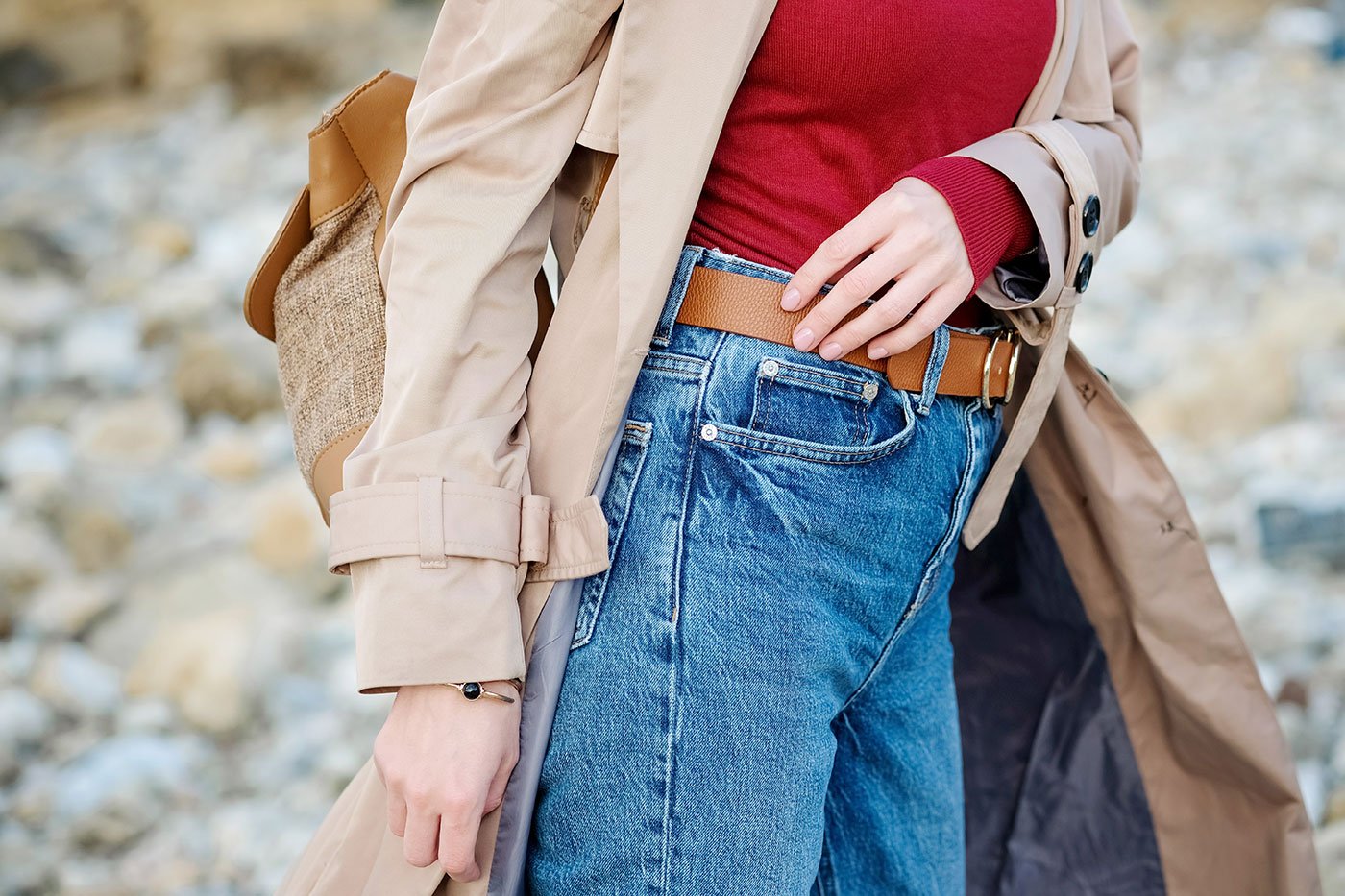 Das Foto die Seitenansicht einer Frau von der Brust bis zum Oberschenkel, die ein rotes Oberteil, eine Jeans und einen beigen Trenchcoat trägt.