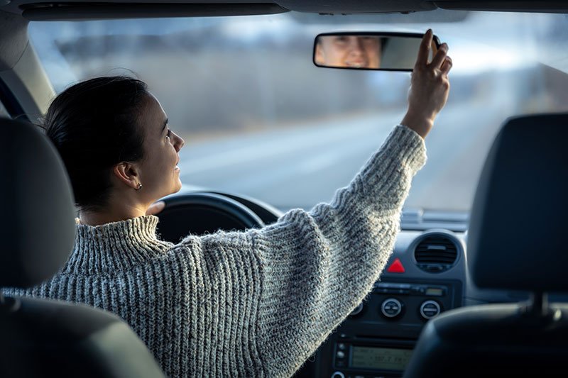 Foto von der Rückbank eines Autos aus in Richtung Windschutzscheibe, auf dem eine Frau auf dem Fahrersitz gerade den Rückspiegel einrichtet.