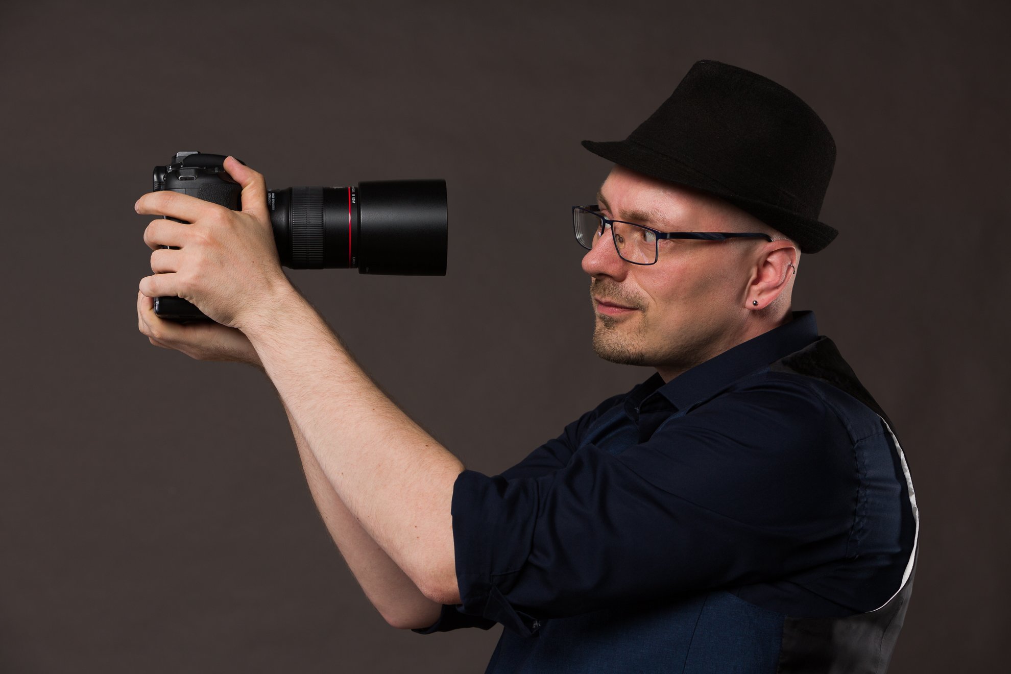 Ein Mann mit Hut und Brille hält eine Kamera mit großem Objektiv in der Hand und schaut direkt in die Linse.