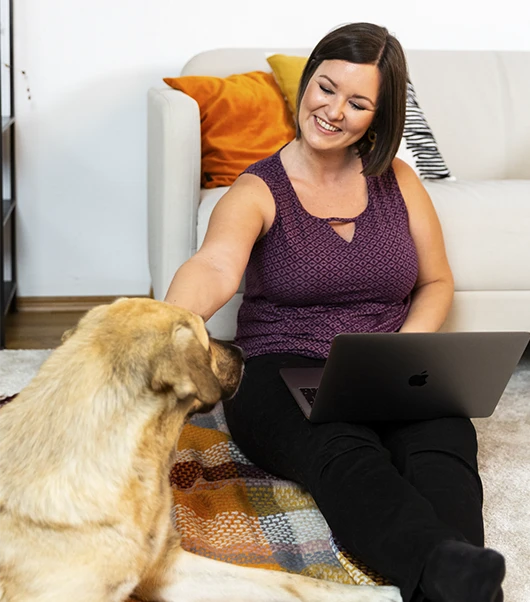 Birgit Spalt-Zoidl sitzt mit ihrem Laptop auf dem Boden und streichelt ihren Hund, der auf einer bunten Decke vor ihr liegt.