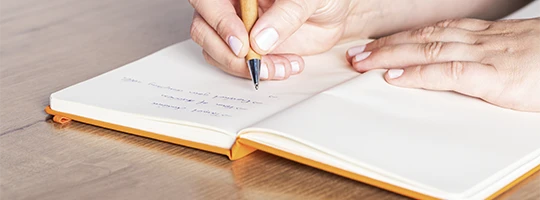 Nahaufnahme einer Hand, die in ein oranges Notizbuch schreibt.