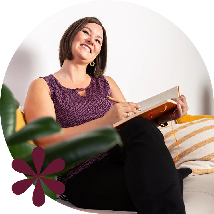 Birgit Spalt-Zoidl sitzt auf einer Couch und blickt gerade nachdenklich lächelnd nach oben, während sie in ein oranges Notizbuch schreibt.