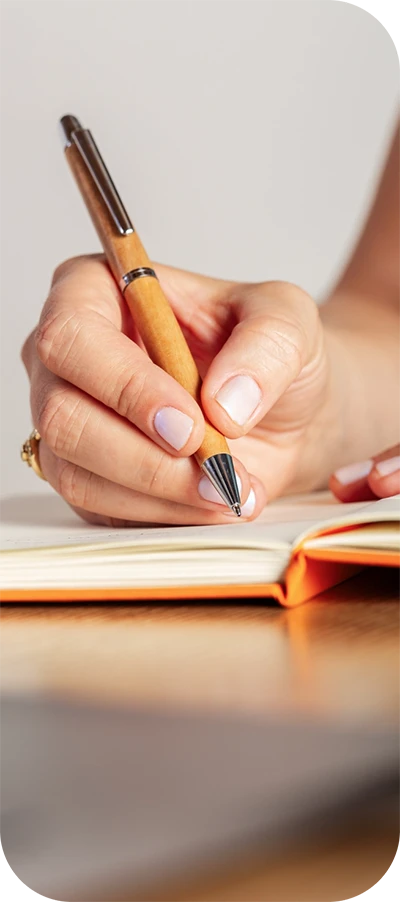 Eine Frauenhand hält einen Holzkugelschreiber und schreibt in ein oranges Notizbuch.