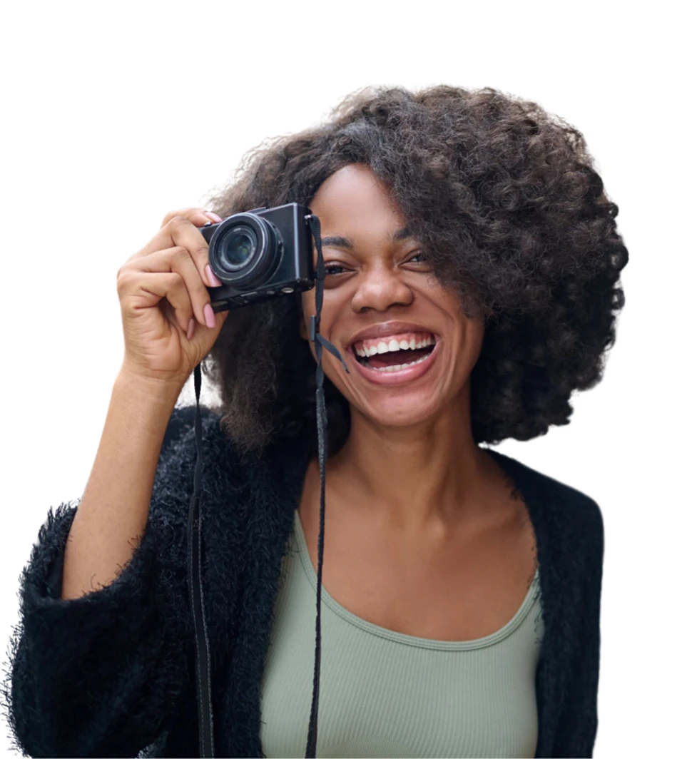 Eine Frau mit schwarzen Locken hält eine Vintage-Kamera hoch und lacht fröhlich in die Kamera.