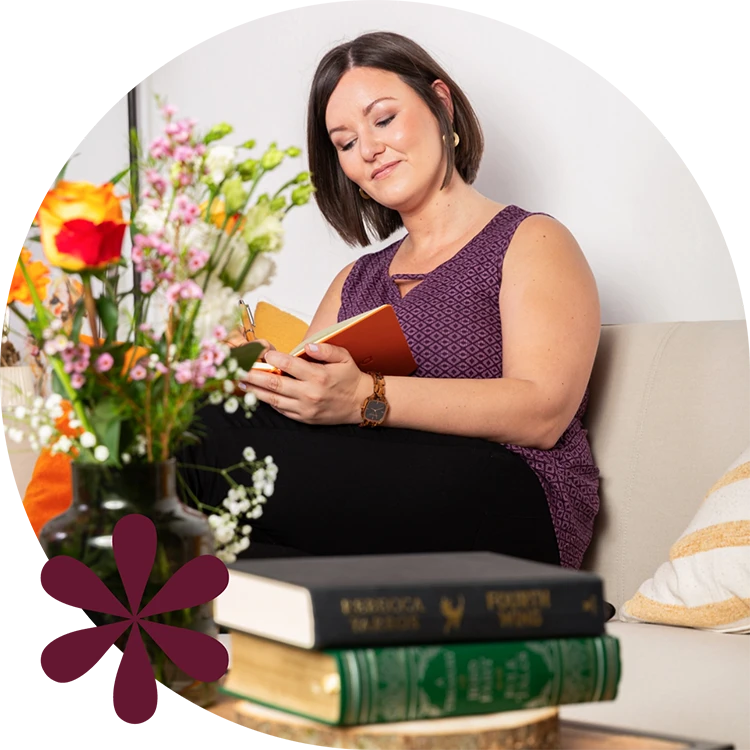 Birgit Spalt-Zoidl sitzt auf einer Couch und schreibt in ein oranges Notizbuch, unscharf im Vordergrund sind auf einem Tisch Blumen und zwei Bücher.